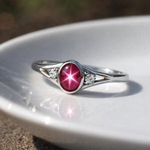 May include: A silver ring featuring an oval-cut, star ruby gemstone. The ruby is a deep red color with a six-ray star effect. Two small, round, clear gemstones flank the ruby on either side. The ring is set on a white surface.