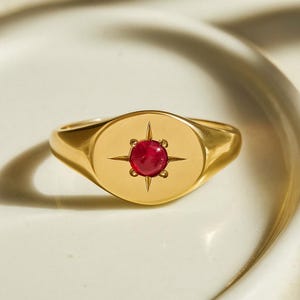 Peut inclure: Une bague chevalière en or avec une face ovale. La face présente une pierre précieuse rouge ronde au centre, entourée d'un motif en forme d'étoile. La bague est posée sur une surface blanche.