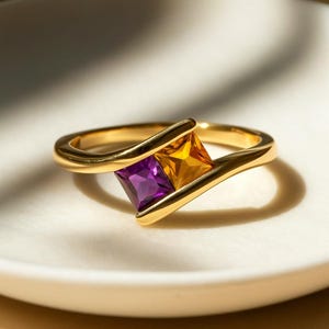 Op de afbeelding: Een gouden ring met twee vierkante edelstenen, een paarse en een gele. De band van de ring buigt om de stenen heen, wat een modern ontwerp creëert. De ring ligt op een wit oppervlak.