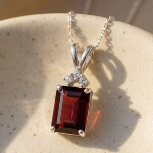 Puede incluir: Un collar con colgante de plata con una piedra preciosa rectangular de color rojo intenso. La piedra preciosa está acentuada con tres pequeñas piedras transparentes. El collar se muestra sobre una superficie de tono neutro.