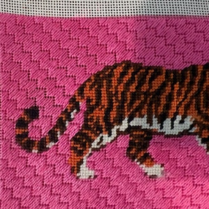 Pode incluir: Uma tela de ponto de agulha apresentando um tigre com listras laranjas e pretas contra um fundo rosa vibrante. O tigre &eacute; retratado de perfil, mostrando a cauda e as patas traseiras, bordado em um tecido de malha branca com padr&atilde;o texturizado.