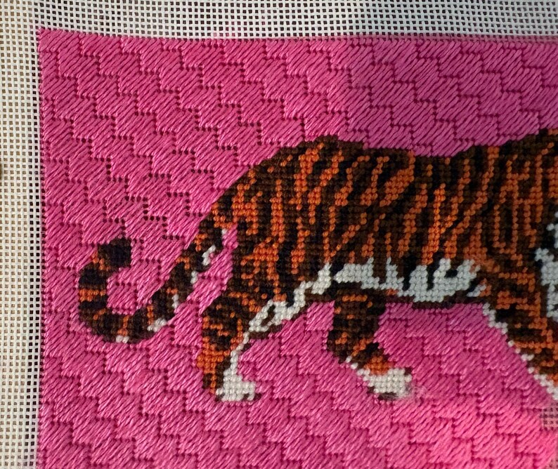 Pode incluir: Uma tela de ponto de agulha apresentando um tigre com listras laranjas e pretas contra um fundo rosa vibrante. O tigre &eacute; retratado de perfil, mostrando a cauda e as patas traseiras, bordado em um tecido de malha branca com padr&atilde;o texturizado.