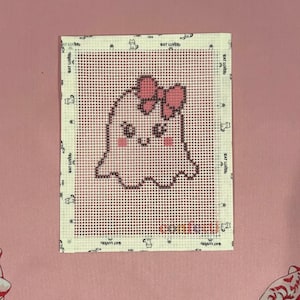May include: A rectangular needlepoint canvas featuring a cross-stitched design of a white ghost with a pink bow on its head, pink cheeks, and dark eyes. The canvas border is printed with the repeating text RAT LOVER and small cat icons.