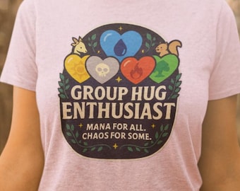 Może przedstawiać: Jasnoróżowa koszulka z napisem "GROUP HUG ENTHUSIAST" i grafiką serc i zwierząt. Grafika zawiera tekst "MANA FOR ALL. CHAOS FOR SOME." Koszulka wykonana jest z miękkiego materiału.