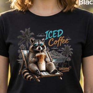 Może przedstawiać: Czarny t-shirt z grafiką przedstawiającą szopa pracza w okularach przeciwsłonecznych, siedzącego na leżaku i trzymającego mrożoną kawę. Wzór zawiera palmy, górski krajobraz oraz napis ICED Coffee i a little bit of SUNSHINE.