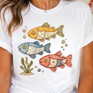 May include: A white t-shirt featuring a graphic print of three fish in yellow, blue, and red. Each fish has a button for an eye and a patchwork-style design with visible stitching. Small bubbles and a green seaweed plant are also printed.