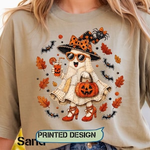 May include: A light beige t-shirt featuring a graphic of a ghost wearing a polka-dot witch hat, orange sunglasses, and red high heels. The ghost holds a jack-o'-lantern bucket and a drink, surrounded by orange autumn leaves, bats, and a spider.