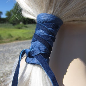 May include: A blue leather ponytail holder with a braided design. The holder is tied around a ponytail of white hair.