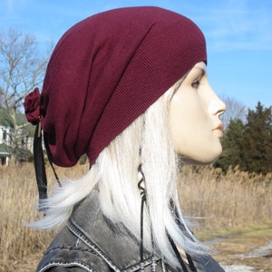 May include: A burgundy slouchy beanie with a black ribbon and a flower detail. The beanie is worn on a mannequin with long white hair.