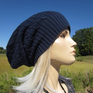 Puede incluir: Un gorro azul marino holgado con un patr&oacute;n de punto de cable. El gorro lo lleva una persona con el pelo largo y rubio y un collar negro con un colgante de plata.
