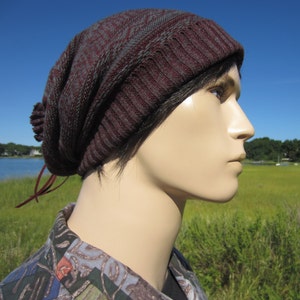May include: A maroon and gray knit beanie hat with a ribbed design. The hat has a slouchy fit and a small pom-pom detail on the side. The hat is worn by a mannequin with a blue sky and green landscape in the background.
