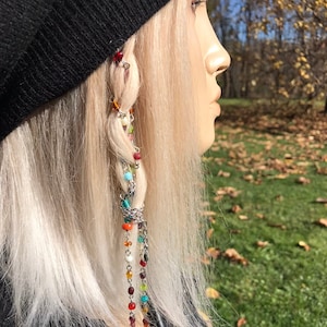 May include: A braided hairstyle with a colorful beaded hair chain. The chain features a variety of beads in different colors, including red, orange, yellow, green, blue, purple, and white. The chain is attached to the braid with a silver clip.