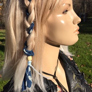 May include: A blonde woman with a blue and white patterned fabric tied into her braid with a light brown wooden bead. She is wearing a black leather jacket with floral embroidery.