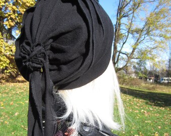 May include: A black knit beanie with a long, flowing tail. The beanie is folded and gathered at the top, creating a unique and stylish look. The beanie is worn by a person with long, white hair.