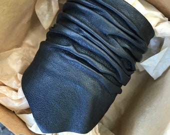Puede incluir: Un brazalete de cuero negro con textura arrugada. El brazalete está en una caja de cartón marrón con papel de seda.