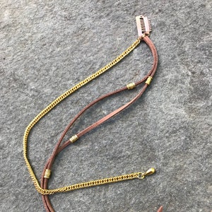 May include: A brown leather cord necklace with gold chain and beads. The necklace has a gold clasp and a gold hair clip at the top.