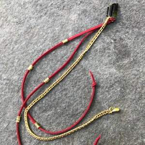 May include: Red suede hair accessory with gold chain and accents. The hair clip features a black metal clip at the top, with a gold chain and red suede strands. The ends of the suede strands are knotted.