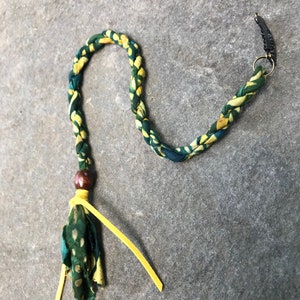 May include: A green and yellow braided hair tie with a wooden bead and a yellow tassel. The hair tie has a metal clip on one end.
