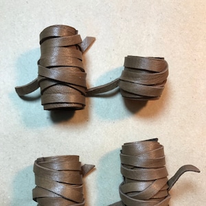 May include: Four rolls of brown leather cord, each tied in a different way. The cords are all the same width and thickness.