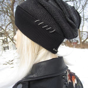 May include: A dark gray beanie with a black ribbed brim and decorative stitching. The hat has a textured knit pattern and a contrasting patterned section. The beanie is worn with a black leather jacket.