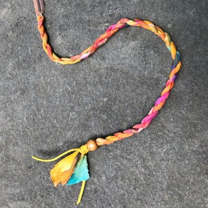 May include: A braided fabric cord with a wooden bead and a colorful fabric tassel. The cord is a mix of orange, yellow, pink, and blue.