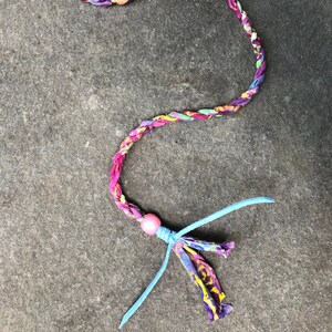 May include: A colorful braided fabric hair tie with a pink bead and a blue tassel. The hair tie has a metal clip at the top.