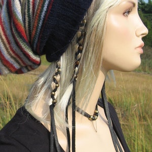 May include: A navy blue and striped knit hat with a pointed top. The hat features vertical stripes in shades of red, brown, tan, and cream. Black leather ties hang from the hat. The mannequin is wearing a black shirt.