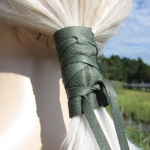 May include: A green leather hair tie with a braided design. The hair tie is tied around a ponytail of white hair.