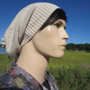 May include: A beige knitted beanie with a long brown cord tie. The beanie is worn by a person with black hair and a light complexion. The person is looking off to the side.