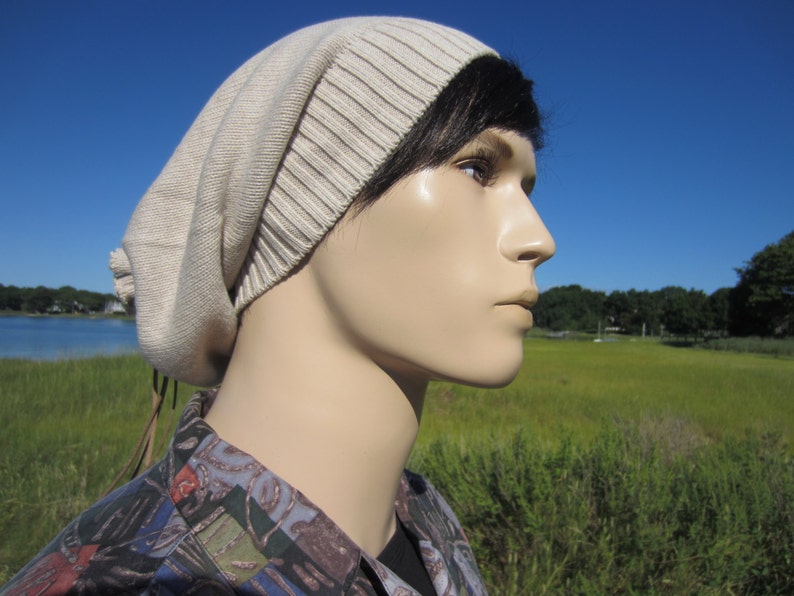 May include: A beige knitted beanie with a long brown cord tie. The beanie is worn by a person with black hair and a light complexion. The person is looking off to the side.