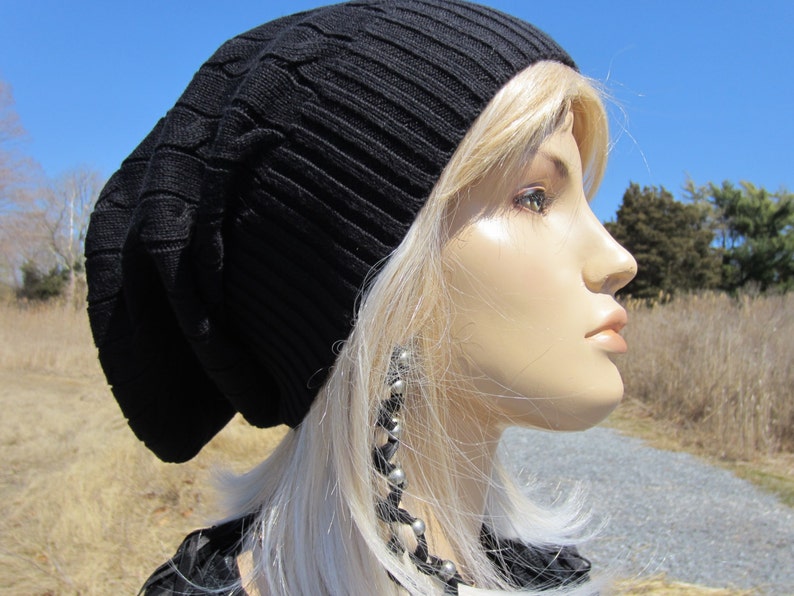 Puede incluir: Un gorro negro de punto con un patr&oacute;n de trenza. El gorro lo lleva una persona con el pelo largo y rubio y pendientes de plata.