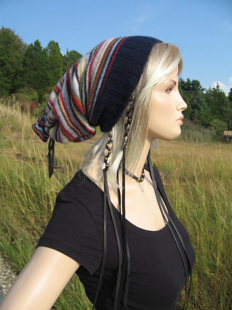 May include: A navy blue and striped knit hat with a pointed top. The hat features vertical stripes in shades of red, brown, tan, and cream. Black leather ties hang from the hat. The mannequin is wearing a black shirt.