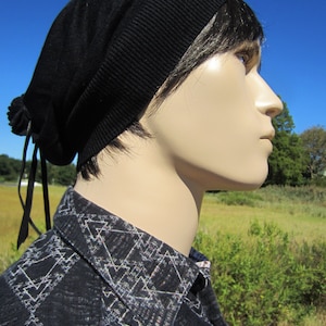 May include: A black slouchy beanie with a long black string tie. The beanie is worn by a mannequin with a black and white geometric patterned shirt.