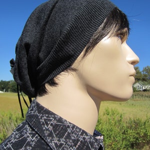 May include: A dark grey slouchy beanie with a long black tie. The beanie is made of a soft, knitted fabric. The person wearing the beanie has dark hair and is wearing a black and white patterned shirt.