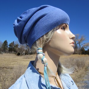 May include: A blue slouchy beanie with a light blue leather hair tie with silver accents. The hair tie is tied around a braid of blonde hair.