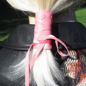 May include: A pink leather ponytail holder with a heart design. The holder is tied around a ponytail of blonde hair.