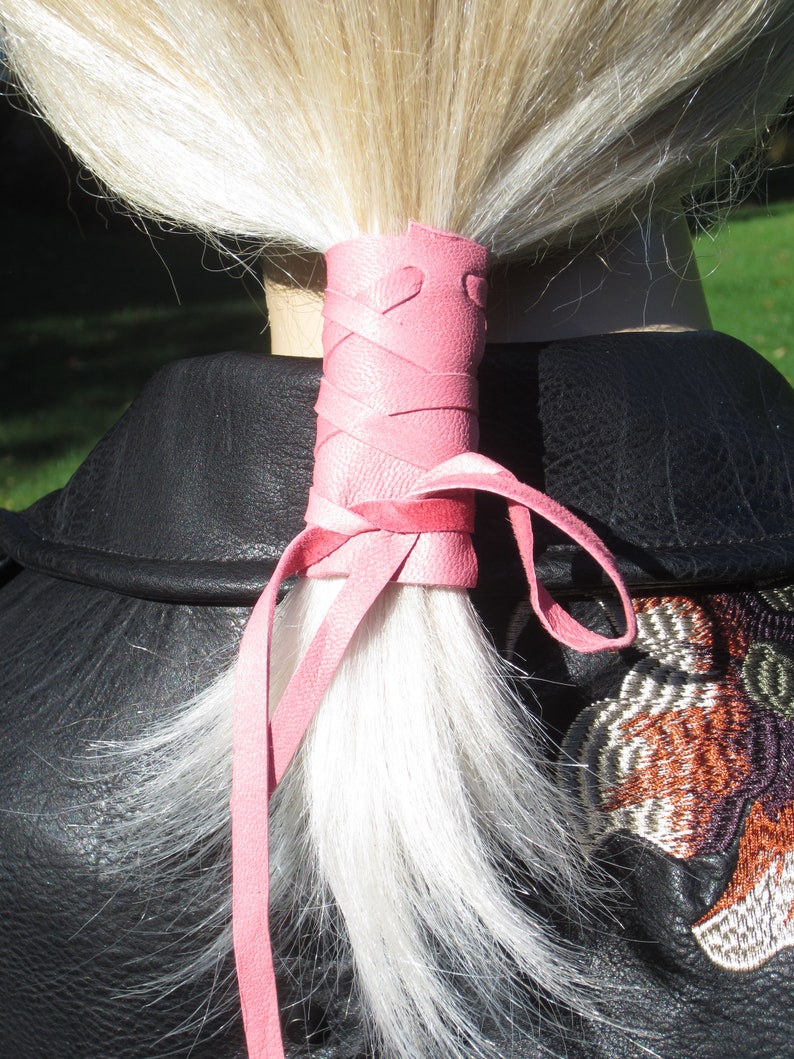 May include: A pink leather ponytail holder with a heart design. The holder is tied around a ponytail of blonde hair.