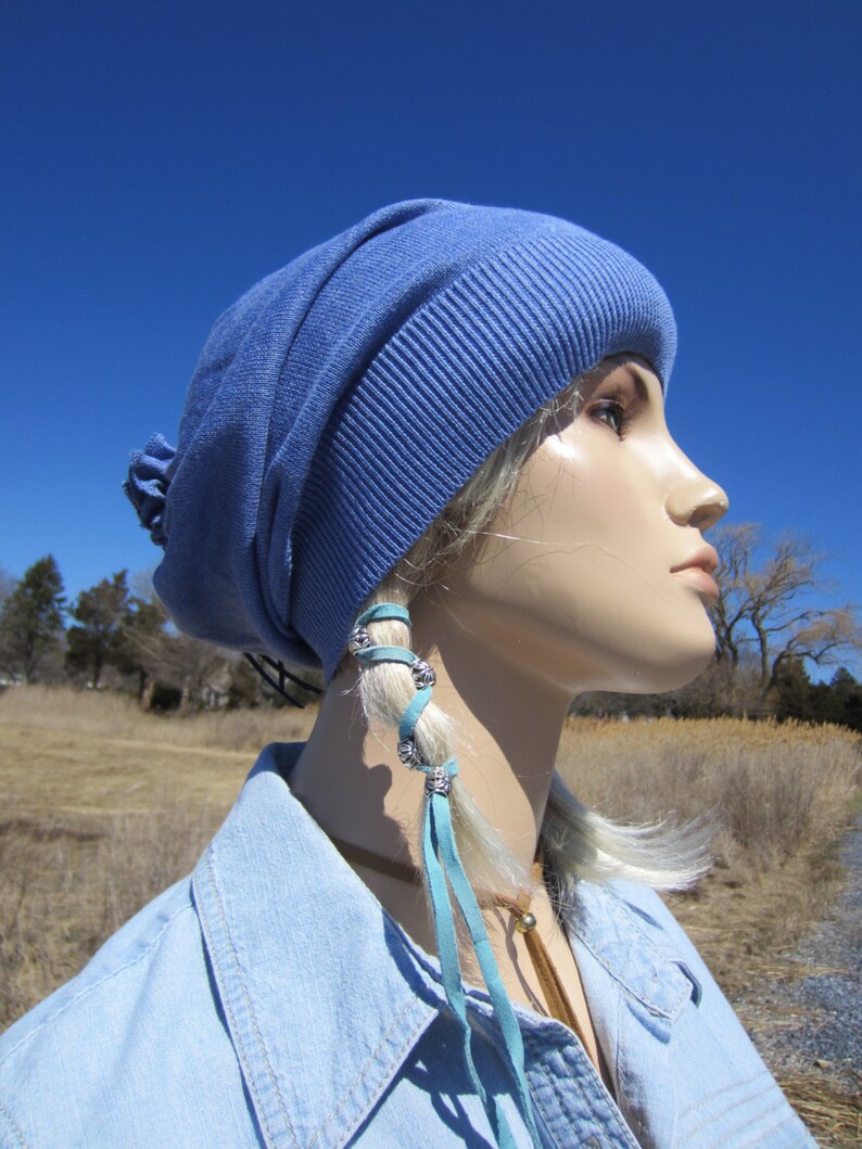 May include: A blue beanie with a light blue leather cord tied around the top. The cord has silver beads and is tied in a knot at the end. The beanie is worn by a mannequin with a blue denim shirt.