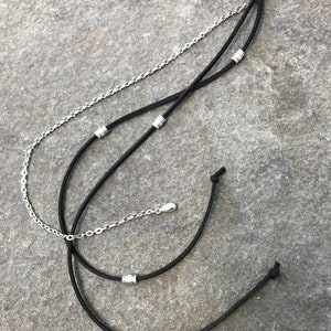 May include: A black hair clip with two black cords and a silver chain attached. The cords and chain have silver beads.