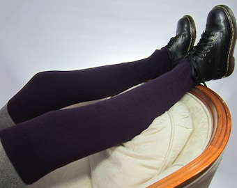 May include: A pair of legs clad in dark purple leggings and black leather boots with laces, resting on a cream-colored upholstered chair. The leggings have a gray cuff at the ankle. The boots are worn and have visible stitching.
