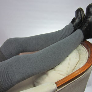 K&ouml;nnte beinhalten: Schwarze Lederstiefel mit Schn&uuml;rung und graue Leggings. Die Leggings sind einfarbig, mit einem dunkleren Graubereich am Knie. Die Stiefel werden beim Entspannen in einem Stuhl getragen.