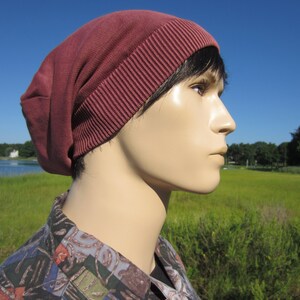 May include: A maroon beanie with a ribbed texture. The beanie is worn on a person's head, with only the side of the face visible.