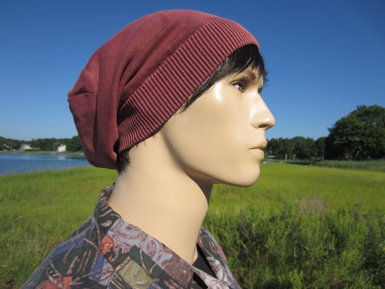 May include: A maroon beanie with a ribbed texture. The beanie is worn on a person's head, with only the side of the face visible.