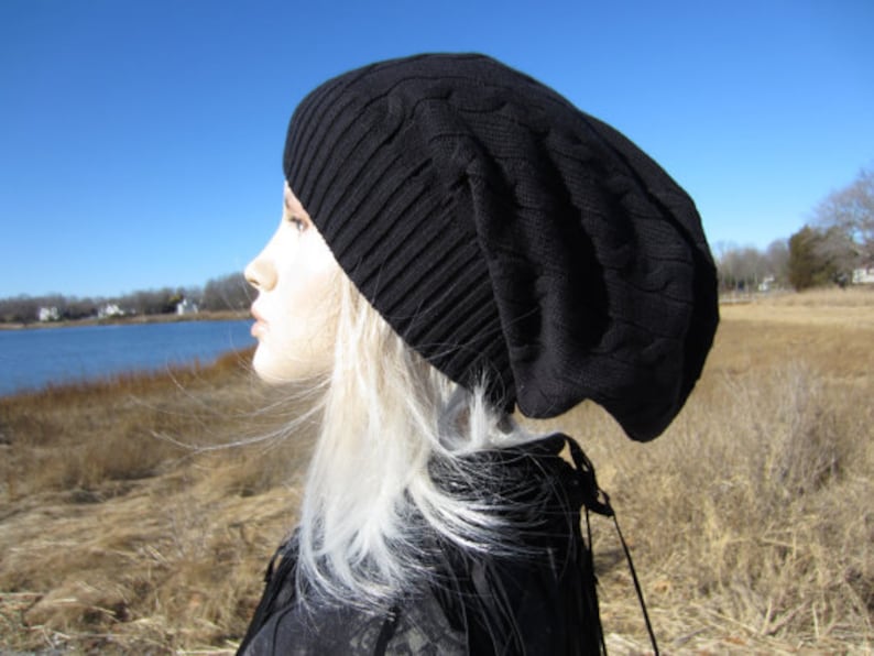 Puede incluir: Un gorro negro de punto de cable con un ajuste holgado. El gorro lo lleva puesto una persona con el pelo largo y blanco. La persona est&aacute; de pie en un campo con hierba alta.