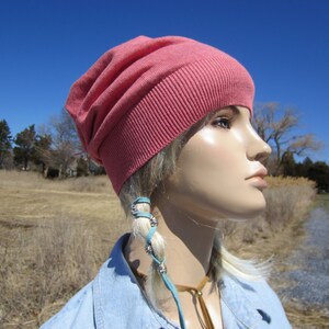 May include: A pink slouchy beanie with a ribbed brim. The beanie is worn by a mannequin with blonde hair and a blue denim shirt. The mannequin is wearing a turquoise leather cord with silver beads around their hair.