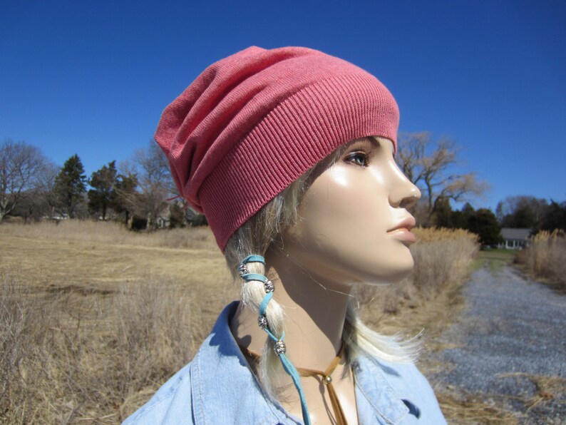 May include: A pink slouchy beanie with a ribbed brim. The beanie is worn by a mannequin with blonde hair and a blue denim shirt. The mannequin is wearing a turquoise leather cord with silver beads around their hair.