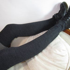 K&ouml;nnte beinhalten: Dunkelgraue Overknee-Beinw&auml;rmer kombiniert mit schwarzen Lederstiefeln. Die Beinw&auml;rmer sind einfarbig, mit einem helleren grauen Abschnitt am Oberschenkel. Die Stiefel haben gelbe N&auml;hte und Schn&uuml;rsenkel.
