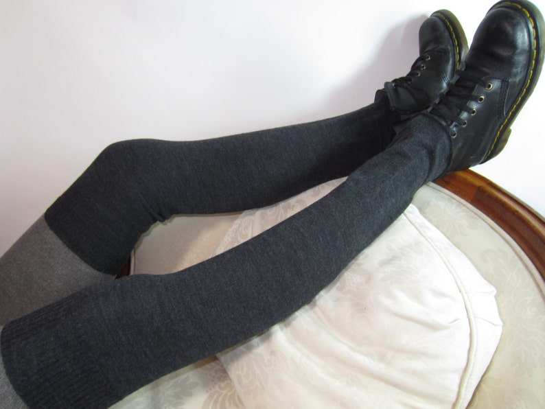 K&ouml;nnte beinhalten: Dunkelgraue Overknee-Beinw&auml;rmer kombiniert mit schwarzen Lederstiefeln. Die Beinw&auml;rmer sind einfarbig, mit einem helleren grauen Abschnitt am Oberschenkel. Die Stiefel haben gelbe N&auml;hte und Schn&uuml;rsenkel.