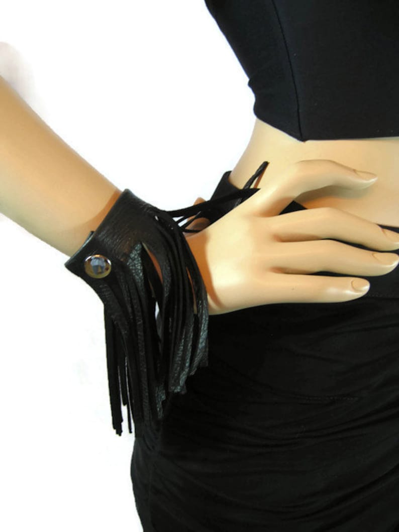 K&ouml;nnte beinhalten: Ein schwarzes Lederarmband mit Fransen. Das Armband hat einen Druckknopfverschluss und einen dekorativen Metallknopf.