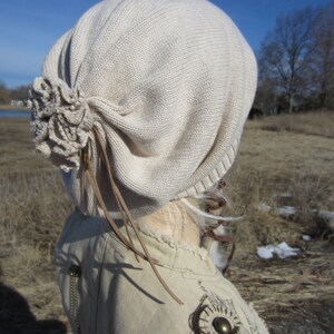May include: A beige knitted beanie with a large flower detail on the side. The beanie has a long brown leather strap that hangs down the back.